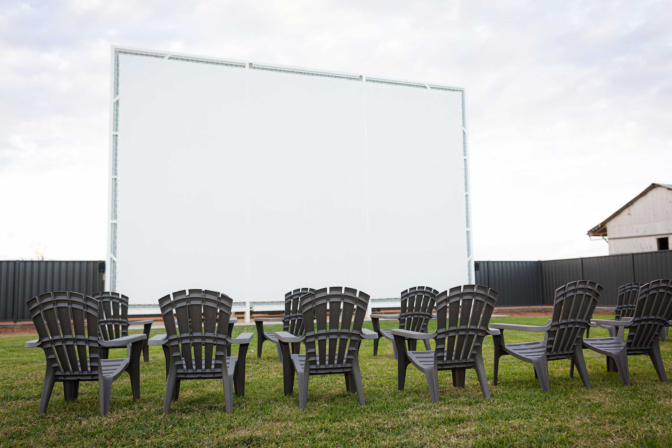 Huge white projector screen sitting on large green lawn with two rows of black lawn chairs facing towards it