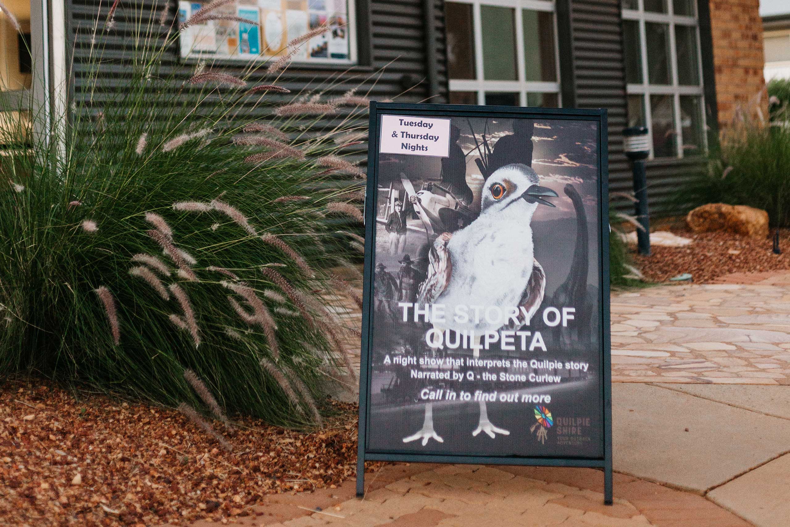 Sandwich board sign with picture of Quilpeta bird and text 'THE STORY OF QUILPETA' outside the entry of black a corrugated iron building and green shrub beside it